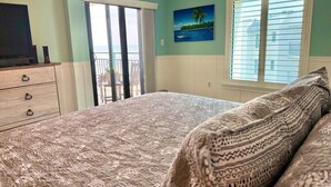 A bedroom with a large bed, a dresser, a painting of a tropical scene, and a view through a sliding door. - Sunny Ormond Beach SOB Condo, Pool, No Drive Beach (Ormond Beach)