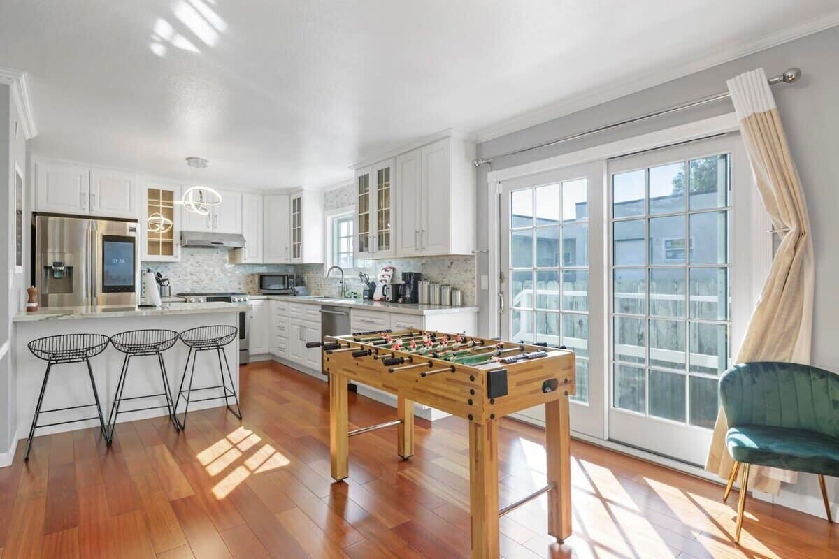 A modern kitchen with a foosball table, bar stools, and a large window with curtains.