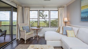 Ein gemütliches Wohnzimmer mit einem weißen Sofa, einem Korbstuhl, einem kleinen runden Tisch und Blick ins Freie durch große Fenster.