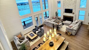 A spacious living room with a large wooden dining table, multiple seating options, and large windows offering views of a snowy landscape. - 6-bedroom cabin-lake access .5 mile-hot tub-sauna-6' spiral slide-Modern Comfort (Island Park)