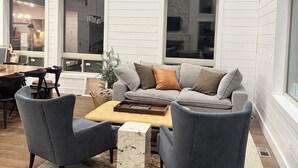 A modern living room with a grey sofa, two armchairs, a coffee table, and a stone accent piece. Large windows provide natural light and views of the outside. - 6-bedroom cabin-lake access .5 mile-hot tub-sauna-6' spiral slide-Modern Comfort (Island Park)