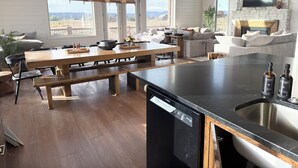 A modern kitchen with a dishwasher, a wooden dining table, and a large window overlooking a scenic view. - 6-bedroom cabin-lake access .5 mile-hot tub-sauna-6' spiral slide-Modern Comfort (Island Park)