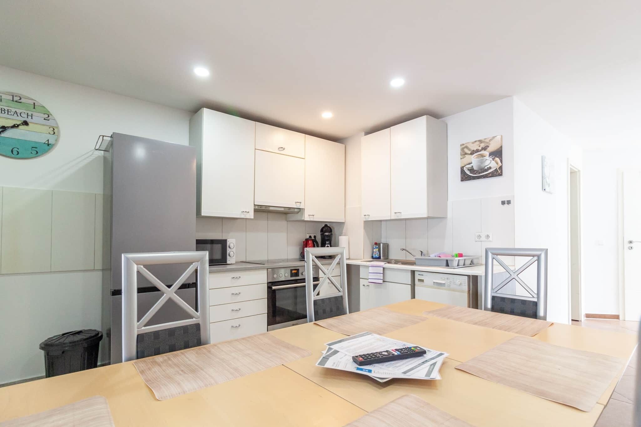 Cozinha moderna com armários brancos, micro-ondas e chaleira. Há uma mesa de jantar com um controle remoto e jornais.