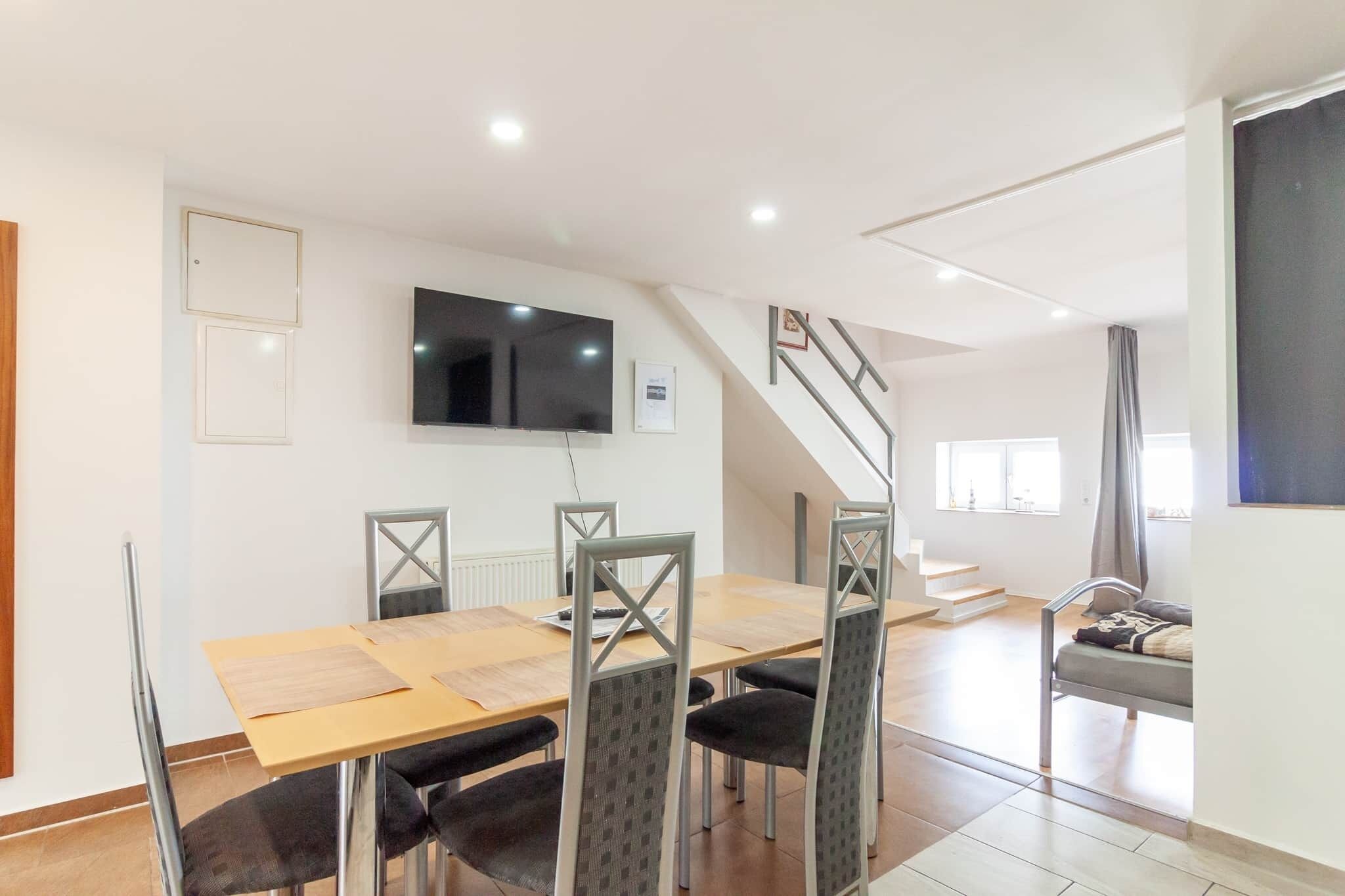 A dining area with a wooden table and chairs, a flat-screen TV mounted on the wall, and a staircase leading to another level.