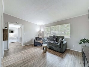 A spacious living room with hardwood floors, a grey sofa, a coffee table, and a large window with blinds. - 3012 E Norfolk Street (Tampa)