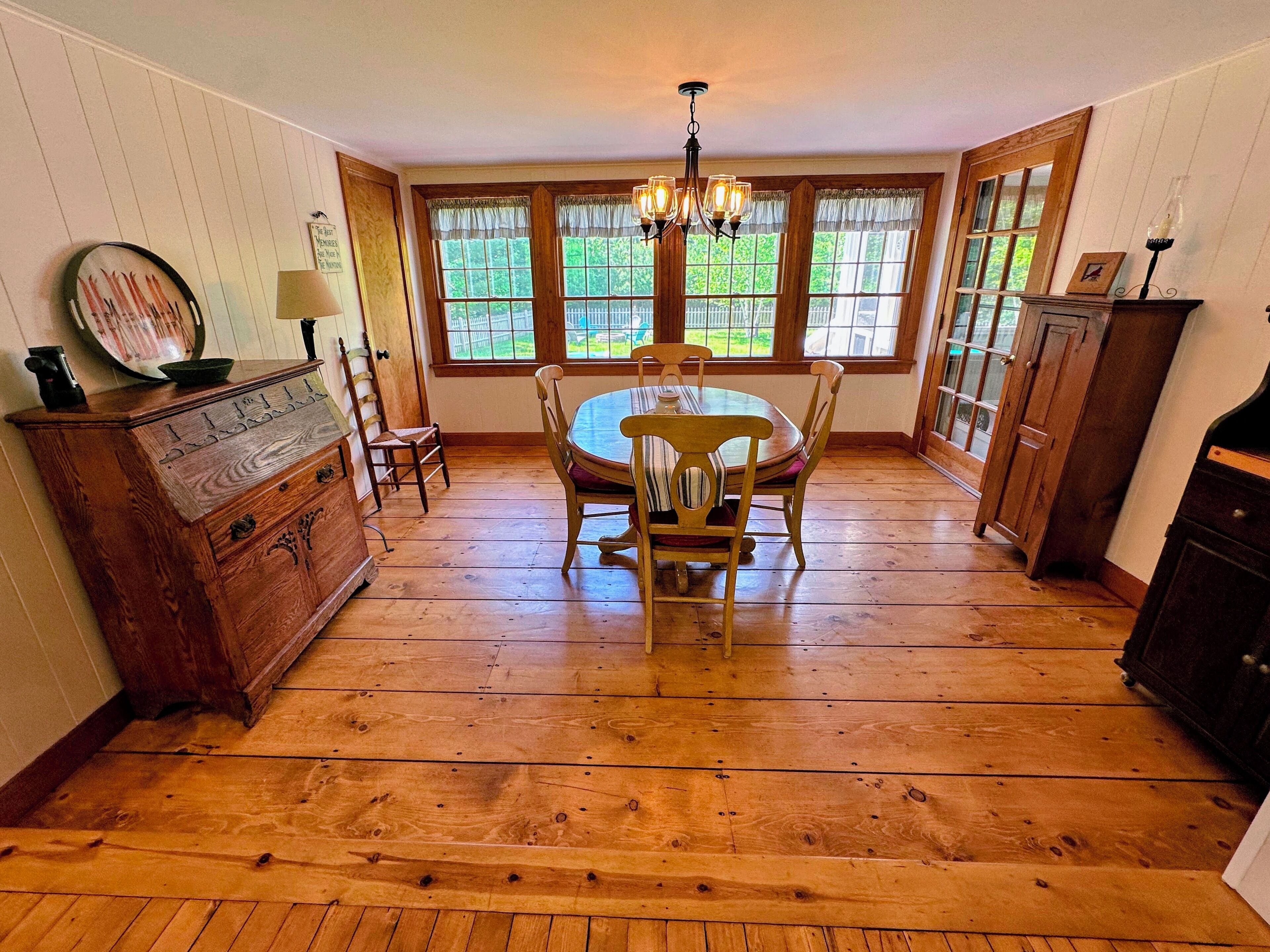 A dining area with a wooden table and chairs, a chandelier, and large windows with a view of greenery.