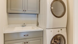 A compact laundry area with a washing machine and dryer stacked on top of each other, a sink, and a cabinet with a drawer below. - 404a The Hilltop House (Virginia Beach)