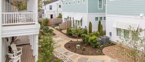 A residential area with a paved walkway, a wooden deck, and a small garden area with shrubs and a tree.