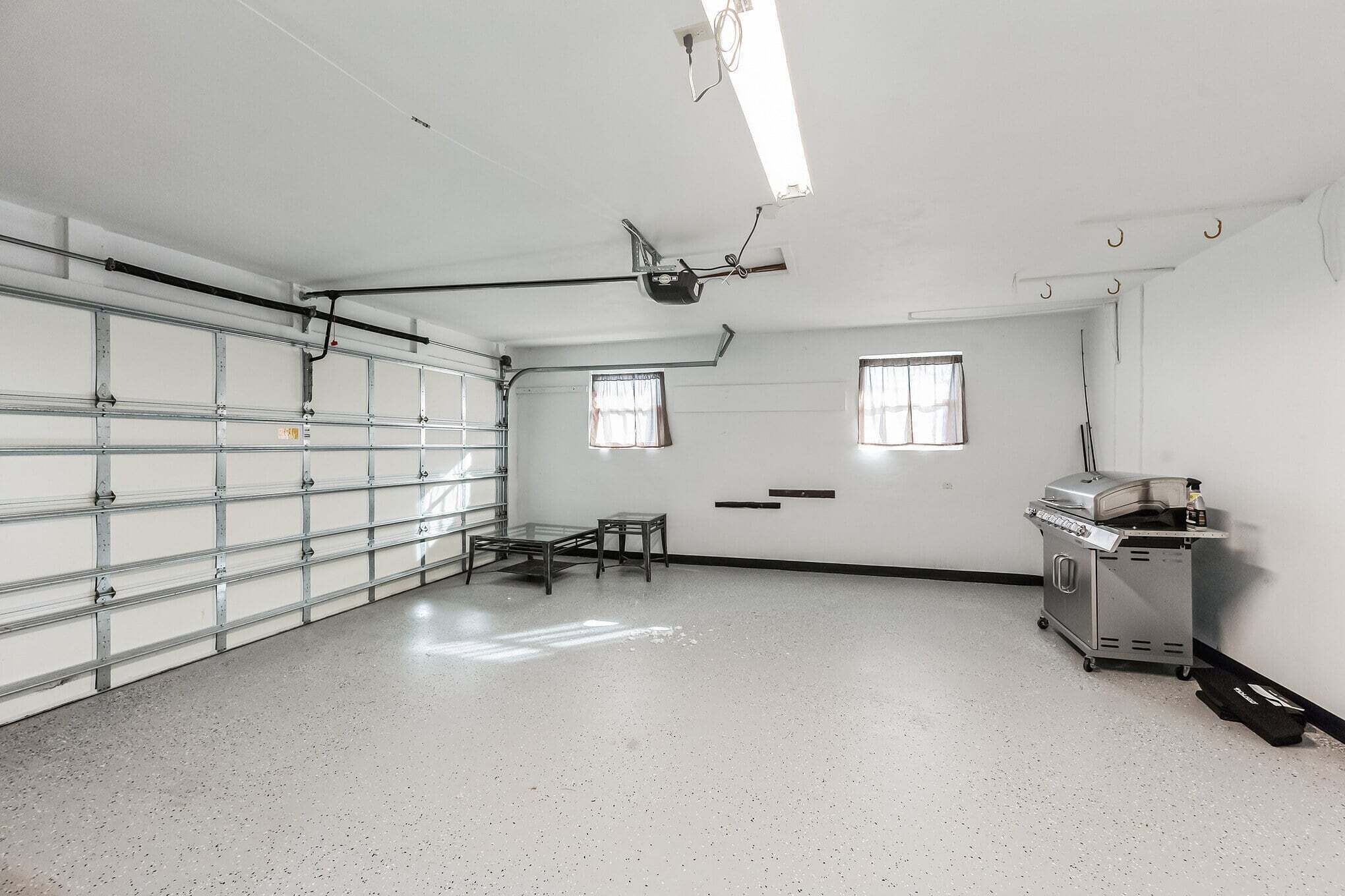 A clean, well-lit garage with a large metal door, a small table, a grill cart, and a ceiling-mounted projector.