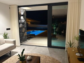A modern living room with a sofa, a coffee table with decorative items, and a view of an outdoor swimming pool at night. - BHF Residences Luxe Family   (Arco da Calheta)