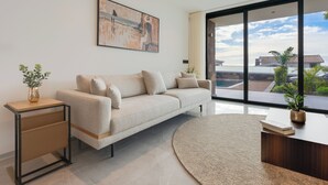 Un salon moderne avec un canapé beige, une table d’appoint sur laquelle est posé un vase, une grande fenêtre donnant sur des immeubles et un tableau accroché au mur.