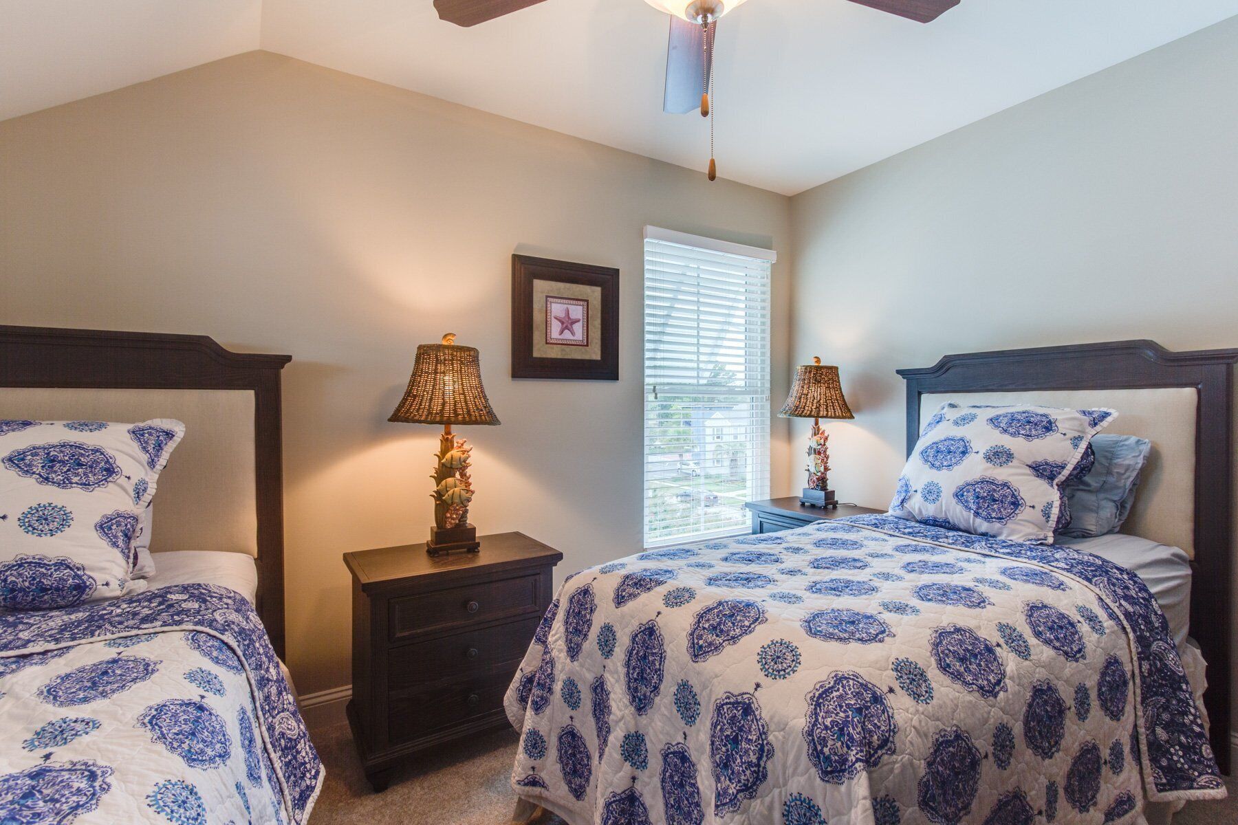 A bedroom with two beds, each with a blue and white patterned bedspread, a nightstand with a lamp, a window with blinds, and a ceiling fan.