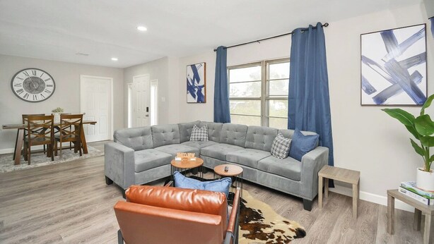 A modern living room with a sectional sofa, a dining table with chairs, a clock, and artwork on the walls. - 5BR 3BA Home With Hot Tub Near Stadiums & Airport (Houston)
