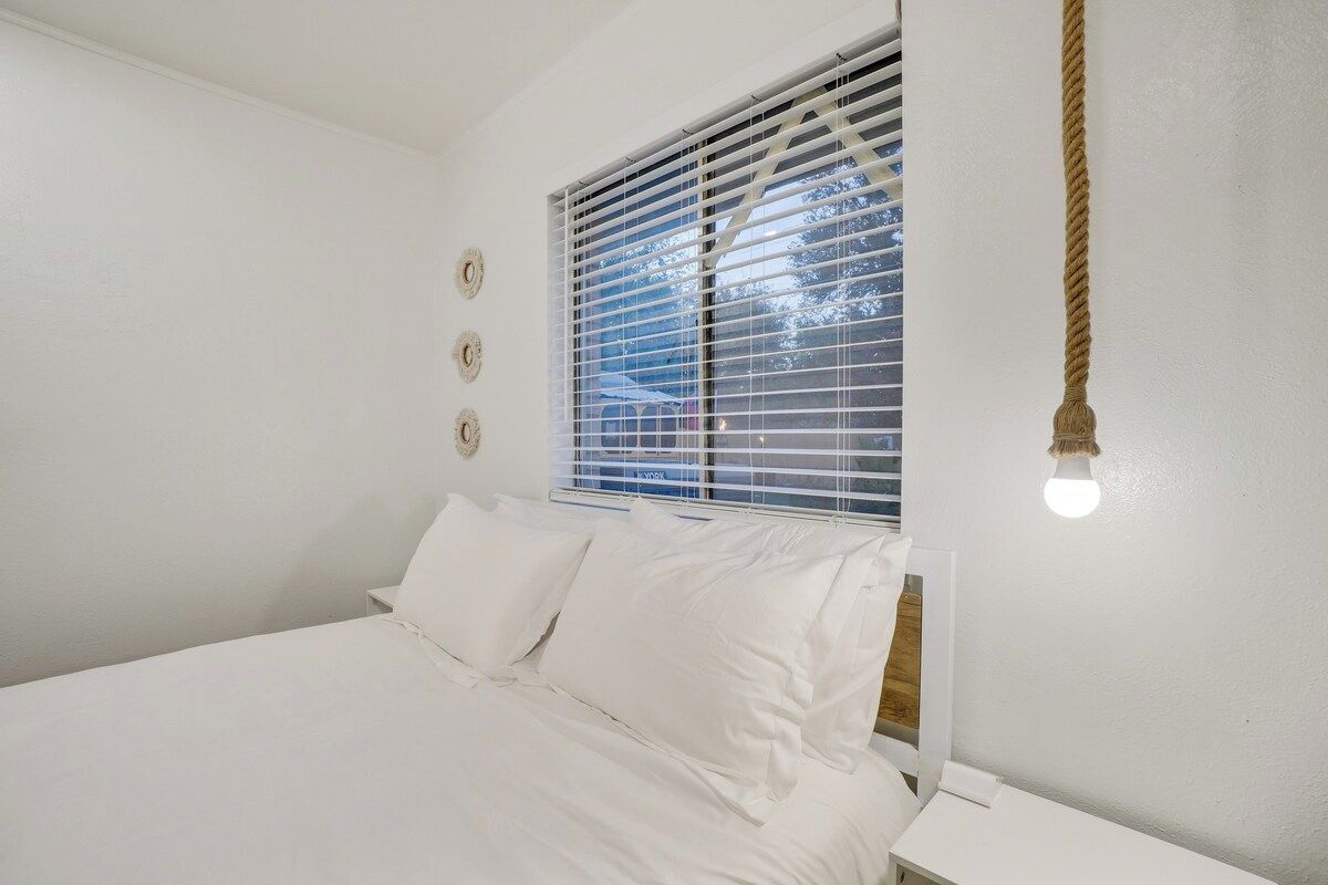 A bedroom with a bed, white walls, a window with blinds, and a hanging light fixture.