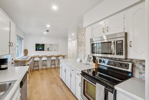 A modern kitchen with white cabinets, a stainless steel oven, a microwave, and a dining area with stools. - 4BR 2B Home With Pool, Hot Tub, Fire Pit (Irving)