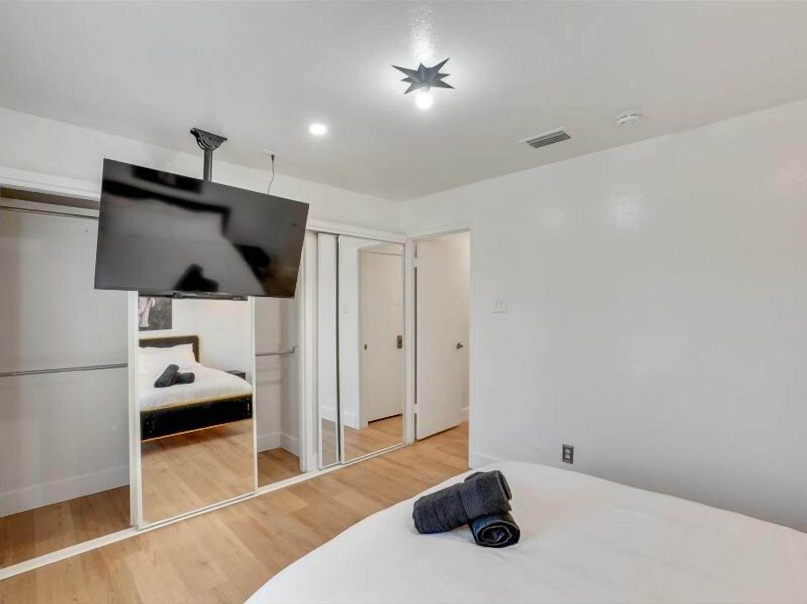 Un dormitorio moderno con un espejo grande, un televisor de pantalla plana montado en la pared, una cama con una toalla enrollada y una puerta que da a otra habitación.