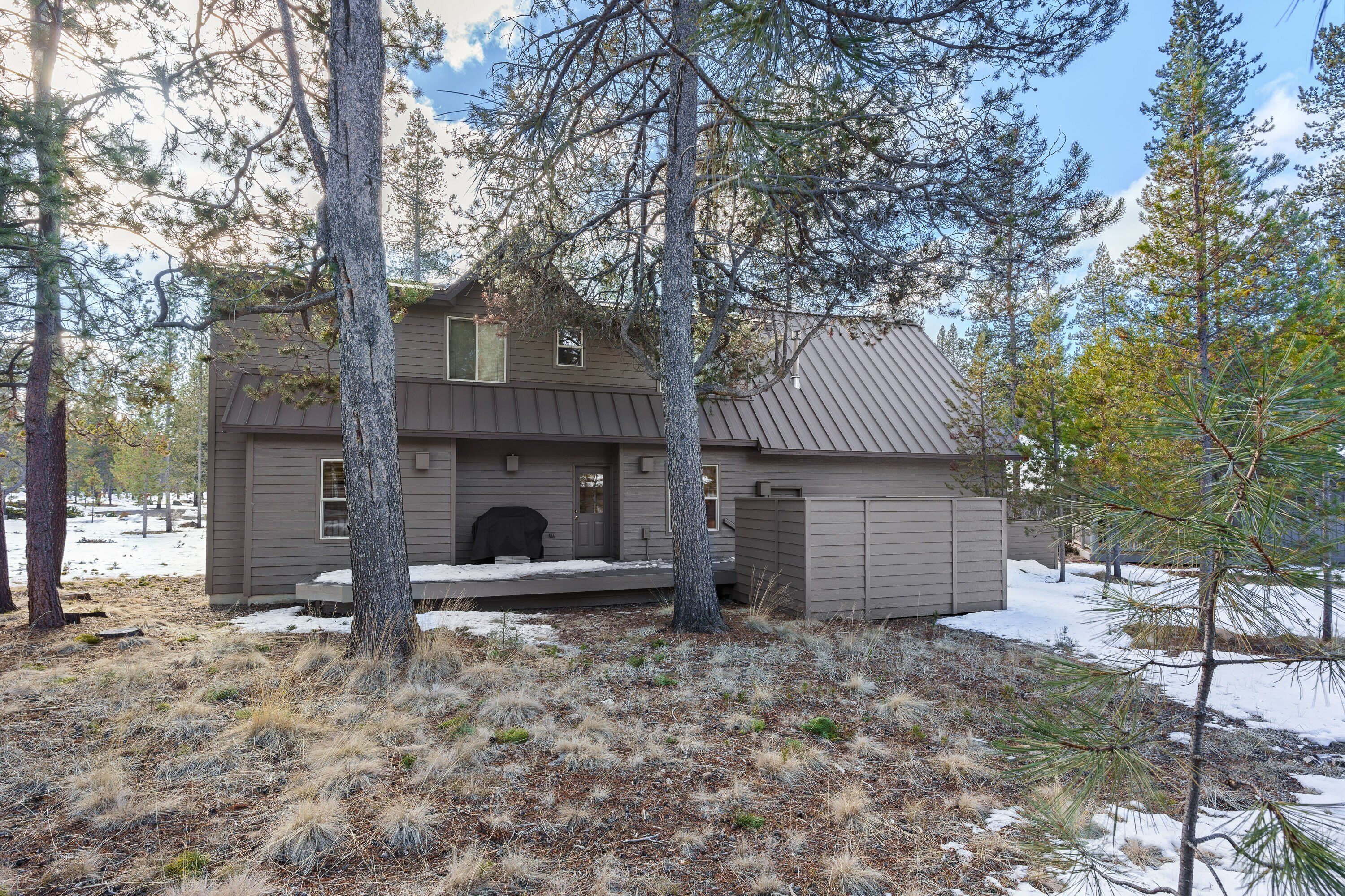 A two-story house with a front porch, surrounded by tall pine trees and a snowy landscape.
