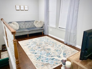 Una sala de estar con un sofá gris, un tapete estampado, una escalera de madera y una televisión.