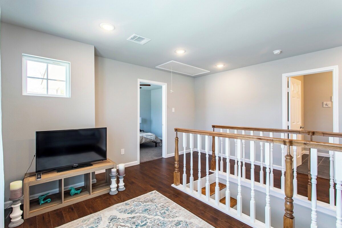 A spacious living area with a flat-screen TV on a wooden stand, a patterned rug, a staircase with white railings, and a hallway leading to another room.