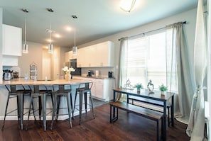 Cocina moderna con una isla central, taburetes de bar, luces colgantes y una mesa de comedor con banco.