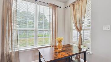 A room with a wooden table, beige curtains, and a window with blinds.