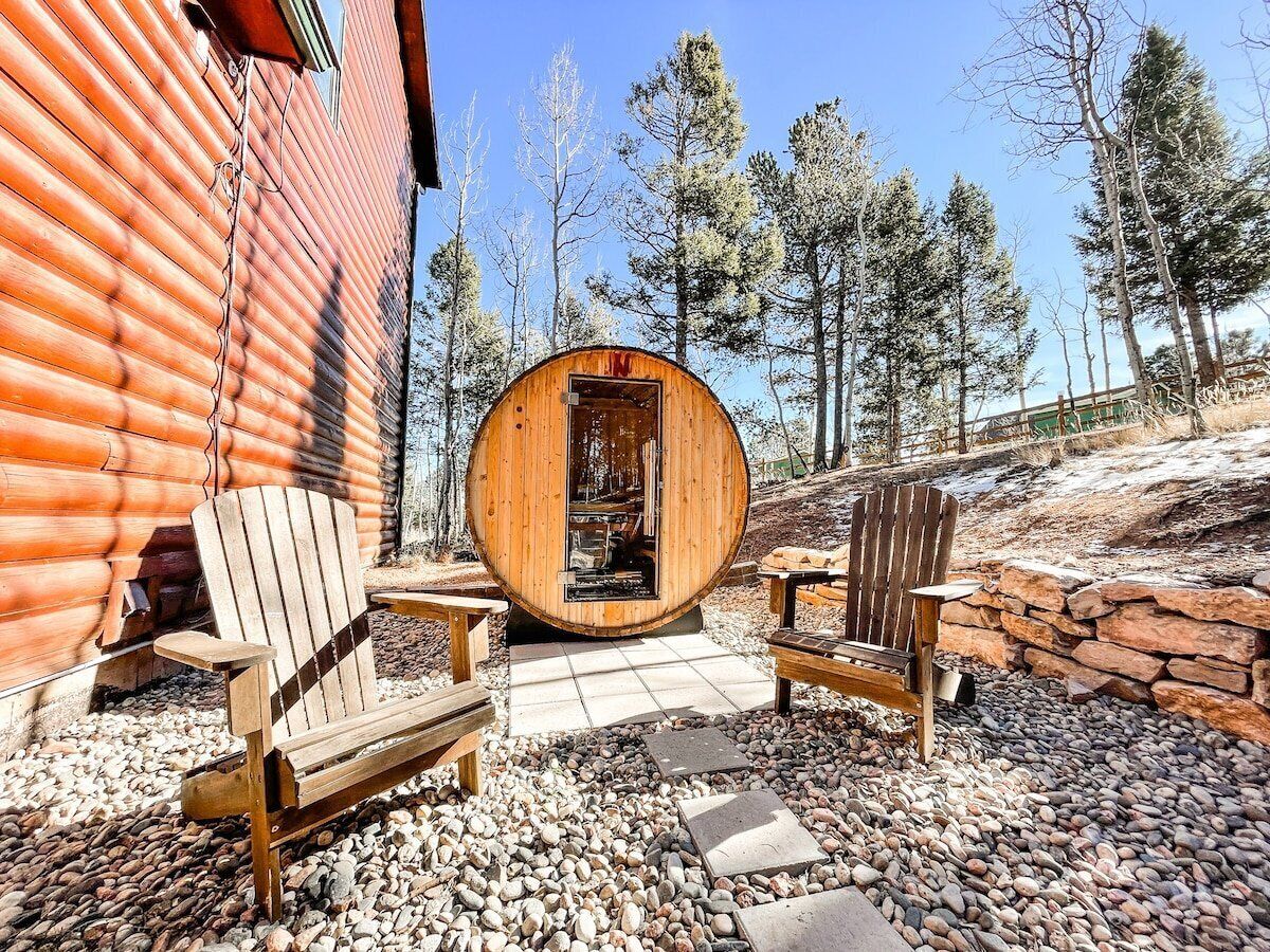 Un sauna en bois doté d’une porte circulaire, de deux fauteuils Adirondack en bois et d’un mur en pierre.