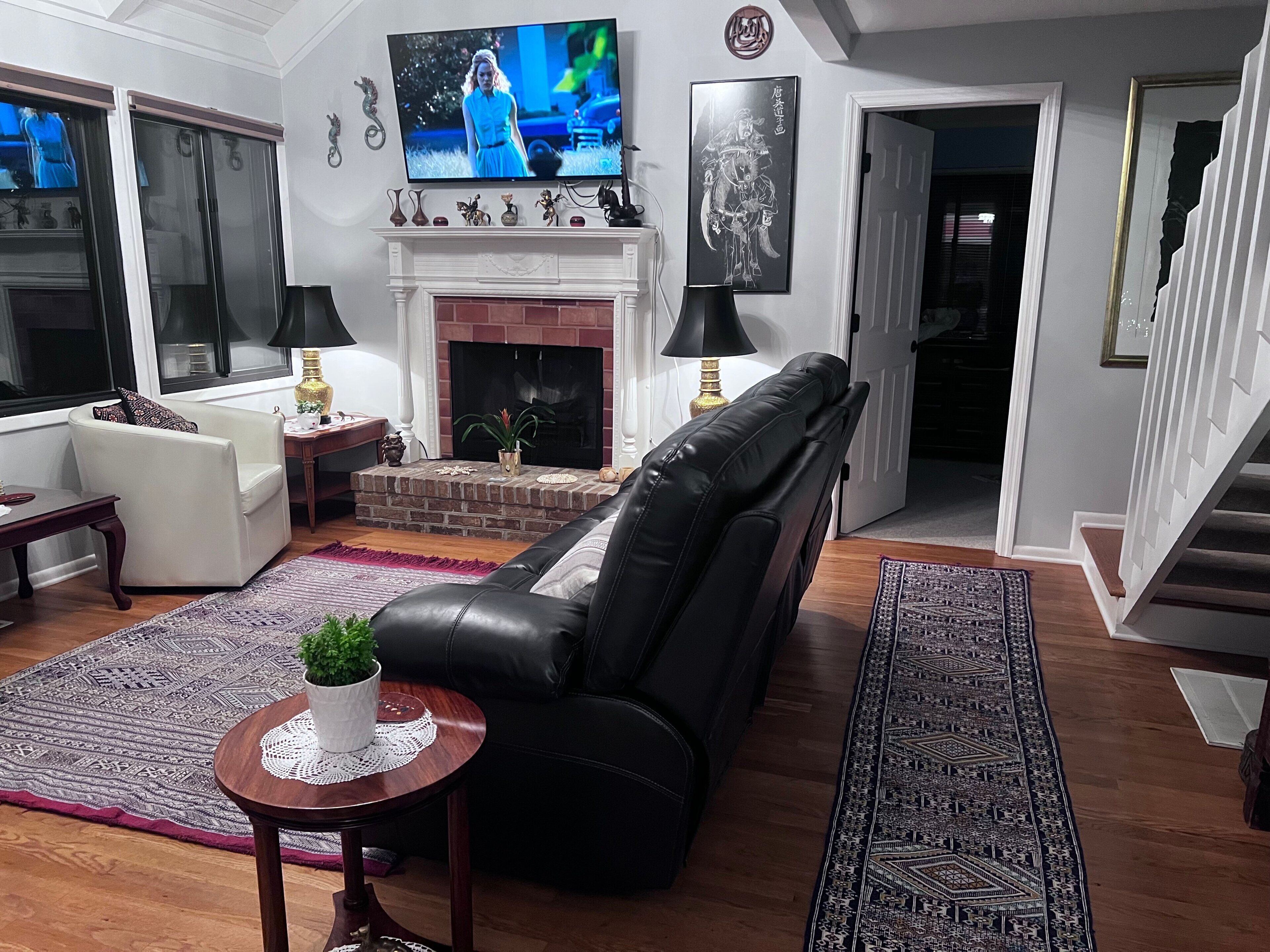 A living room with a fireplace, a black leather sofa, a white armchair, a TV, and a staircase leading to another level.