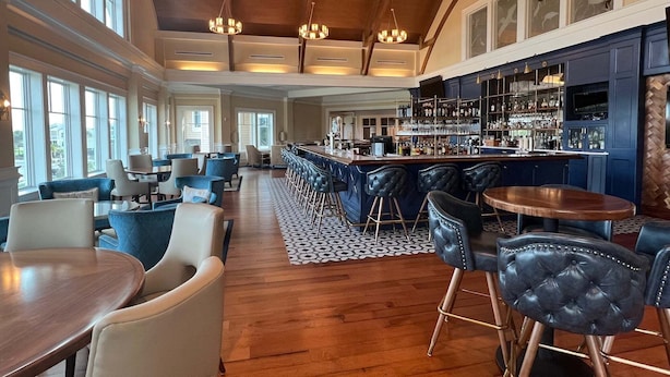 A spacious bar area with high ceilings and large windows, a seating area with sofas and chairs, and a bar counter with shelves of bottles. - 905 Sealoft (Seabrook Island)
