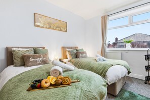 A hotel room with two beds, green bedding, a fruit and donut tray, and a view of a house through the window. - Contractor or Corporate- 2 Bedroom Home | Office| Sleeps 6| Bills Inc (Bolton)