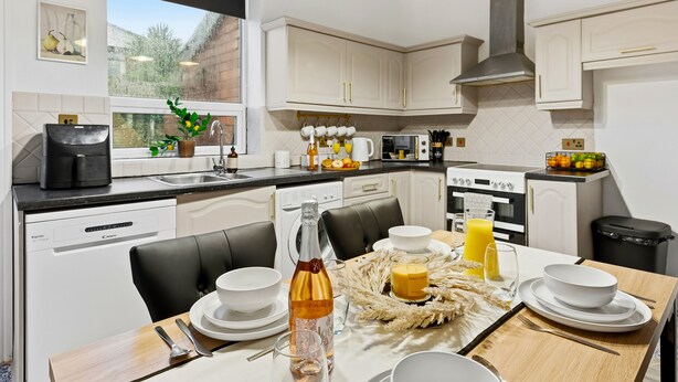 A modern kitchen with white cabinets, a black countertop, and a dining area with a table set for two. - Contractor or Corporate- 2 Bedroom Home | Office| Sleeps 6| Bills Inc (Bolton)