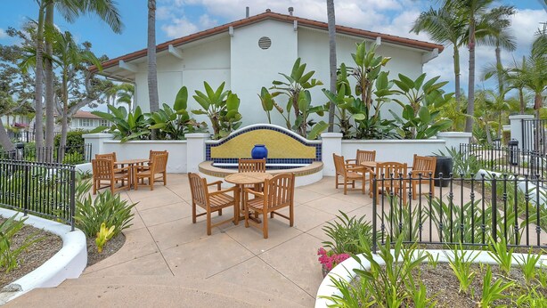 A patio area with wooden tables and chairs, surrounded by palm trees and a white building with a tiled roof.
