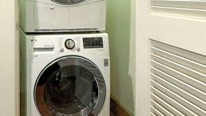 Two stacked front-loading washing machines in a laundry room with a tiled floor and a door with horizontal blinds.