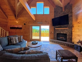 A cozy cabin interior with a stone fireplace, a large TV mounted on the wall, a sofa with cushions, and a view of the mountains through the door.