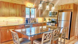 Une cuisine dotée de meubles en bois, d’un îlot en granit, de tabourets de bar, d’appareils électroménagers en acier inoxydable et de luminaires suspendus.