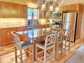 A kitchen with wooden cabinets, a granite island, bar stools, stainless steel appliances, and pendant lighting.