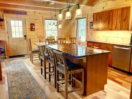 Interior de una cabaña de madera con una gran mesa de comedor, sillas, una zona de cocina y un refrigerador empotrado.