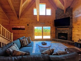 A cozy cabin interior with a wooden ceiling, a stone fireplace, a large sofa, and a view through a glass door.