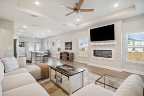 A spacious living room with a large sofa, a coffee table, and a fireplace. There is a kitchen area with a dining table and chairs, and a ceiling fan. - 95070 Gina Street (Fremont)
