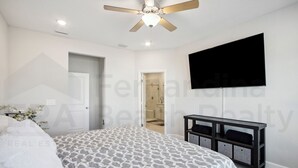 Un dormitorio con cama, ventilador de techo, televisor montado en la pared y puerta que da a un baño.