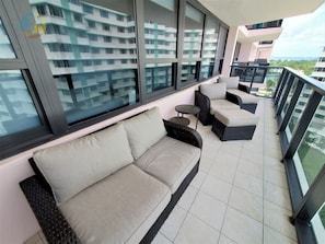 Un balcon avec un canapé et des fauteuils, de grandes fenêtres et une vue sur d’autres immeubles.