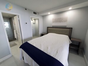Une chambre à coucher comprenant un lit, une table de chevet, une lampe fixée au mur, une porte donnant sur une salle de bain et une petite étagère.