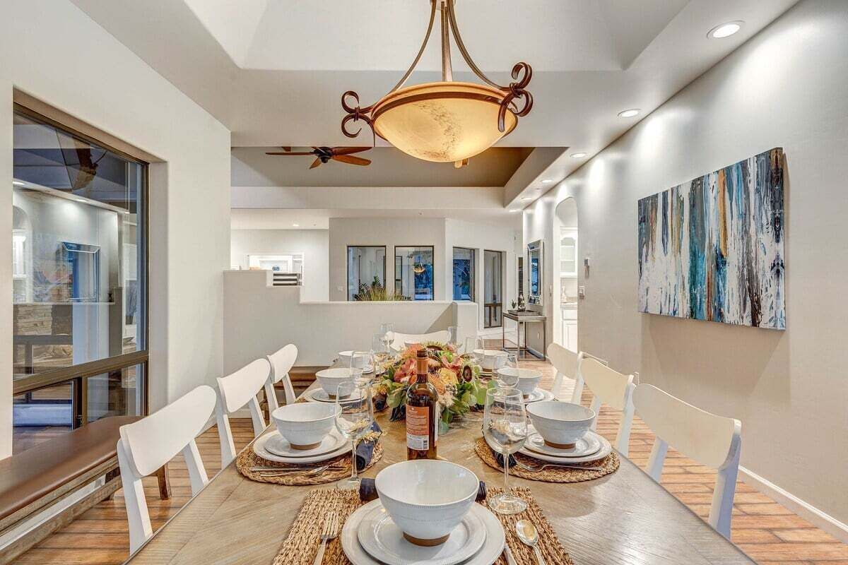 A dining area with a wooden table set for six, white chairs, a chandelier, and artwork on the walls.