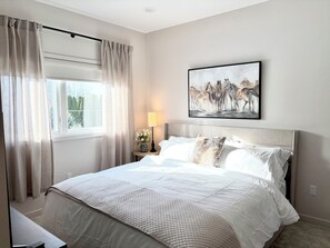 Um quarto com uma cama grande, uma mesa de cabeceira com abajur, uma janela com cortinas e um quadro de cavalos na parede.