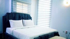Un llit ben fet amb roba de llit blanca i un panell de capçal fosc en una habitació amb parets de blau clar i una finestra amb persianes.