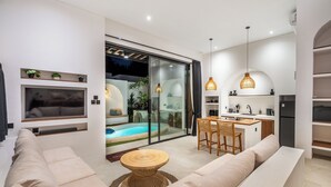 Un saló modern amb un sofà, una zona de menjador amb taula i cadirees, una cuina amb nevera, i una porta de vidre que dona a una piscina.