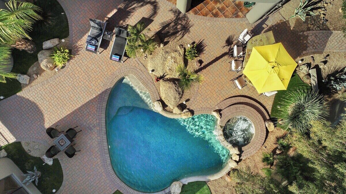 Vista aerea di una zona piscina con piscina circolare, circondata da poltroncine a sdraio e ombrelloni, e diverse piante.