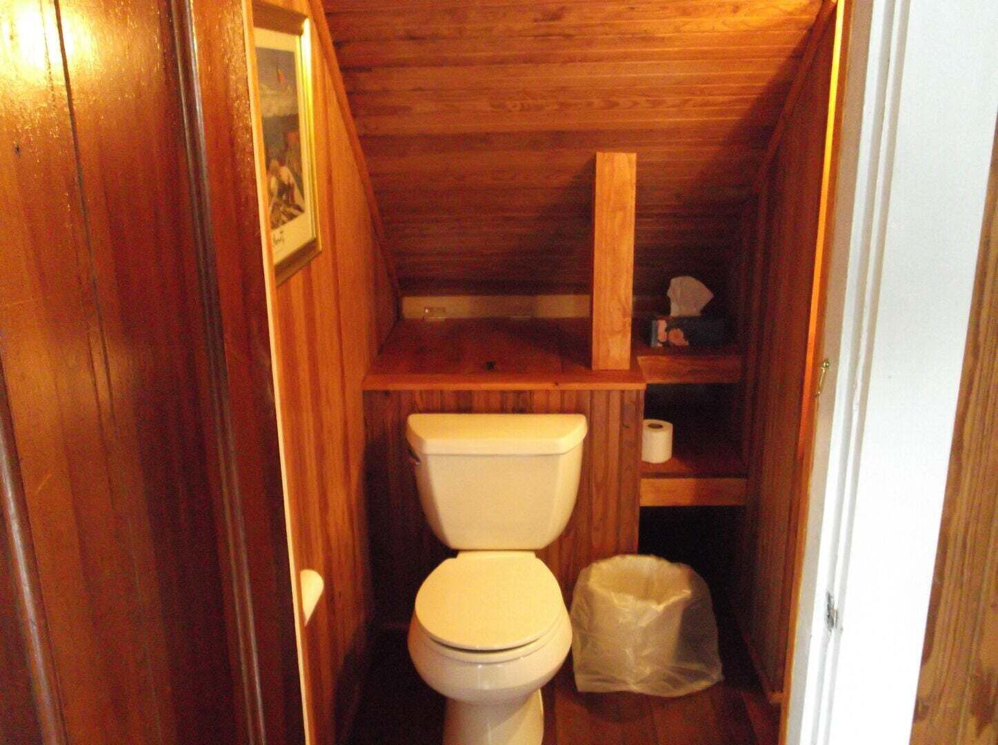 A small, wooden-paneled bathroom with a toilet, a shelf with a roll of toilet paper, and a plastic bag on the floor.