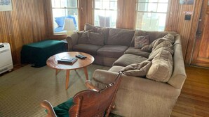 Une pièce aux murs lambrés, avec un canapé d’angle, une table basse ronde en bois et un fauteuil à bascule.