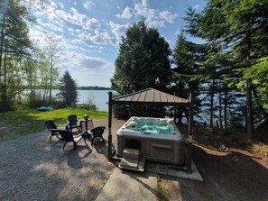 A hot tub under a gazebo by a lake with chairs and a table.