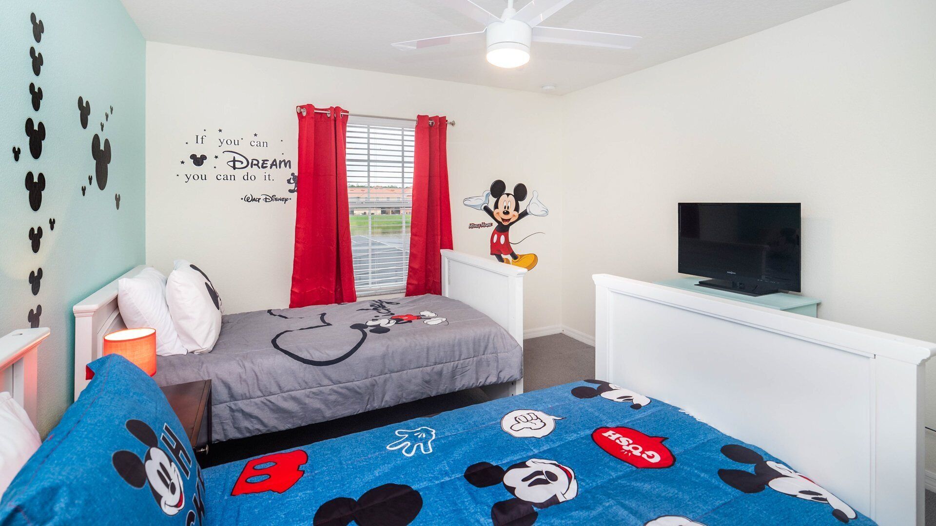 A bedroom with Mickey Mouse wall decals, a bed with a Mickey Mouse-themed blanket, a TV on a white dresser, and a window with red curtains.